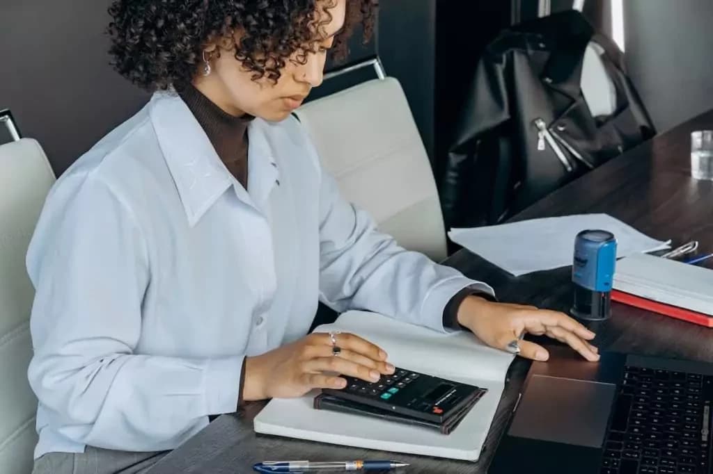 Imagem de uma mulher usando uma camisa social azul claro. Ela está sentada em um escritório e usa um notebook enquanto faz cálculos com uma calculadora. Imagem usada para ilustrar o texto que discute os impostos para o apostador brasileiro