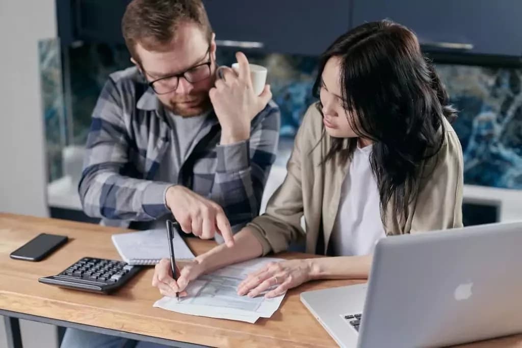 Imagem de um homem e uma mulher em uma mesa. Na mesa vemos vários papéis, que parecem ser contas, além de uma calculadora, um celular e um laptop. Eles estão concentrados fazendo as contas.