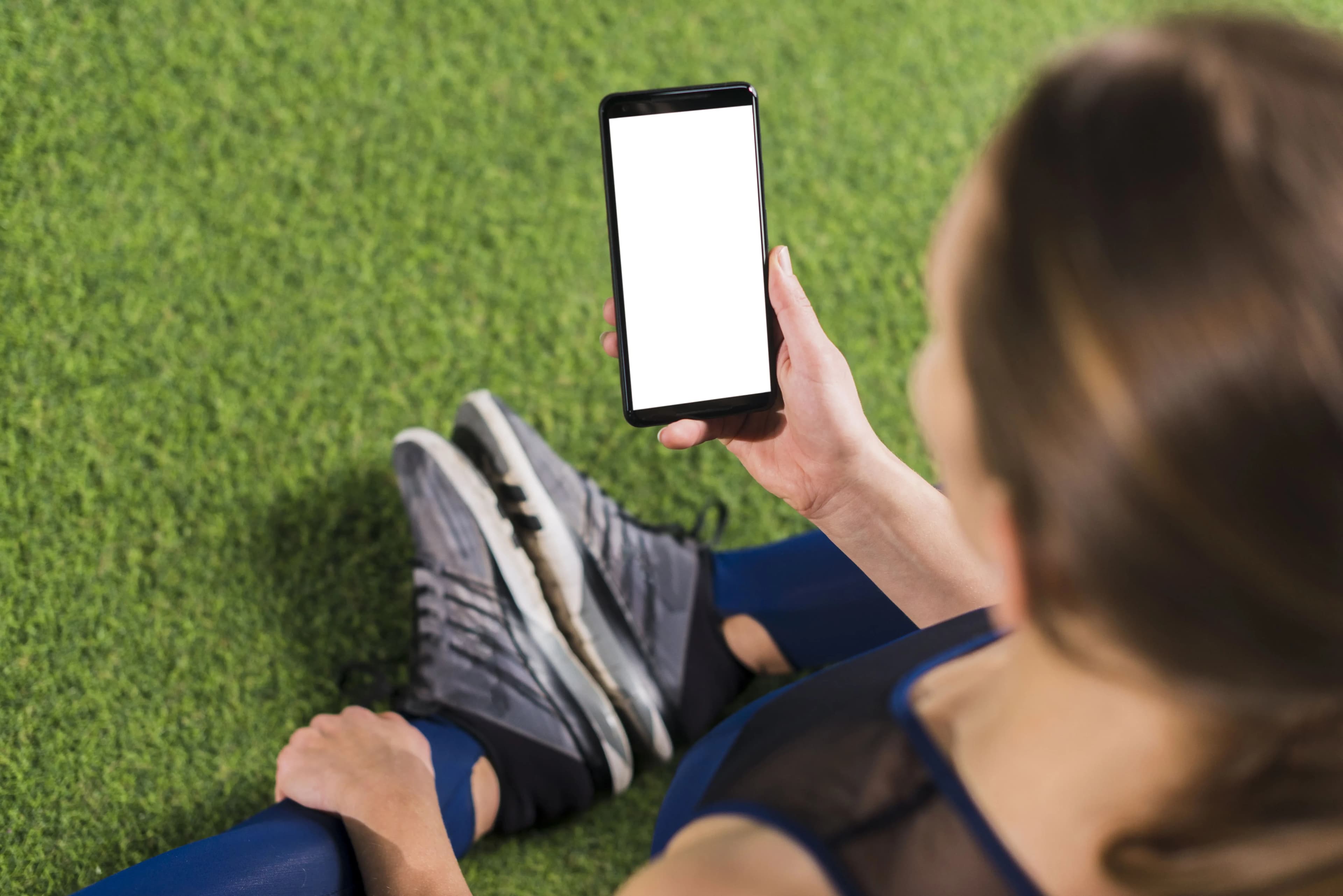 A imagem mostra uma mulher sentada em um campo de futebol olhando para o celular.