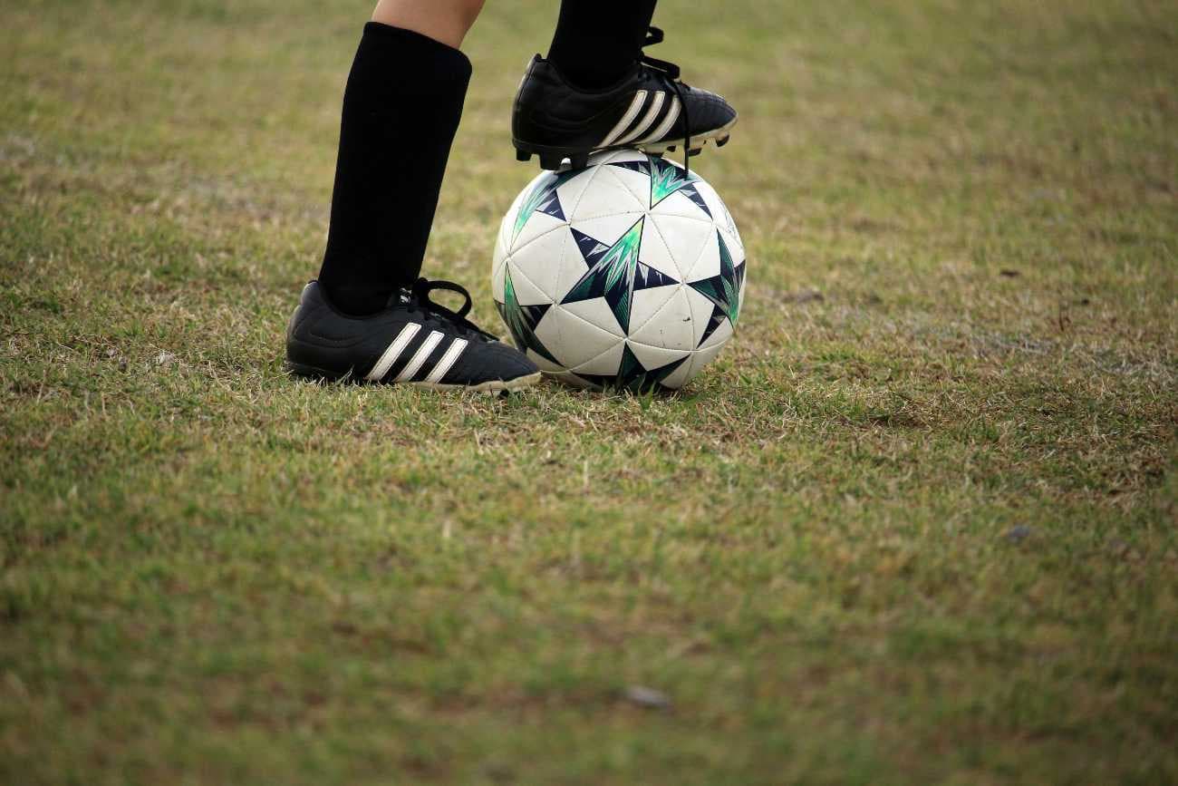 Imagem de uma bola de futebol sendo controlada pelos pés de um atleta, que usa chuteiras e meias pretas.