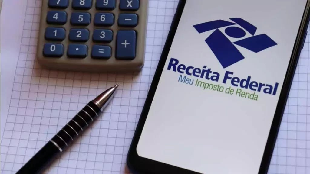 Imagem de um celular com o app do Imposto de Renda aberto, junto vemos uma calculadora e uma caneta