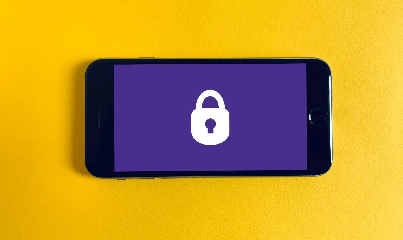 Imagem de um celular com um cadeado na tela, representando a segurança em transações digitais. O celular está sob um fundo amarelo