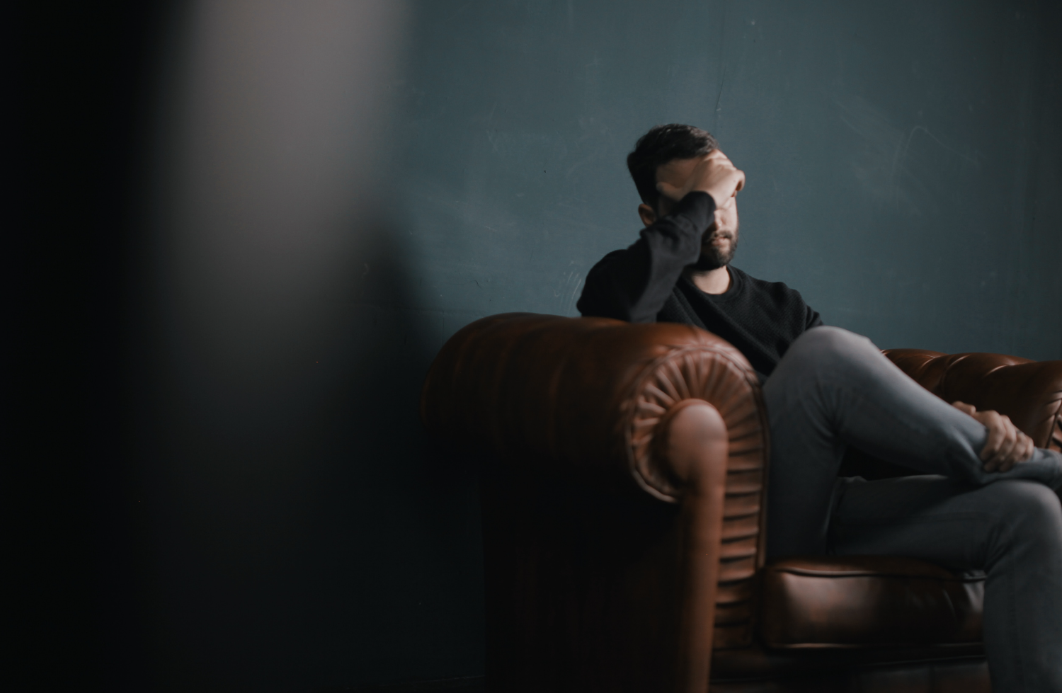 Imagem de um homem sentado em um sofá, usando uma blusa preta e calça cinza. Ele está preocupado, é o que indica a sua expressão facial