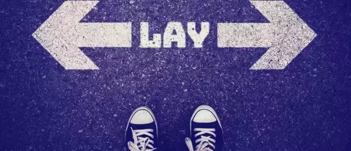 Imagem de uma rua, com o escrito LAY e duas setas, cada uma apontada para um lado. Vemos também parte dos pés de uma pessoa, que usa tênis all star