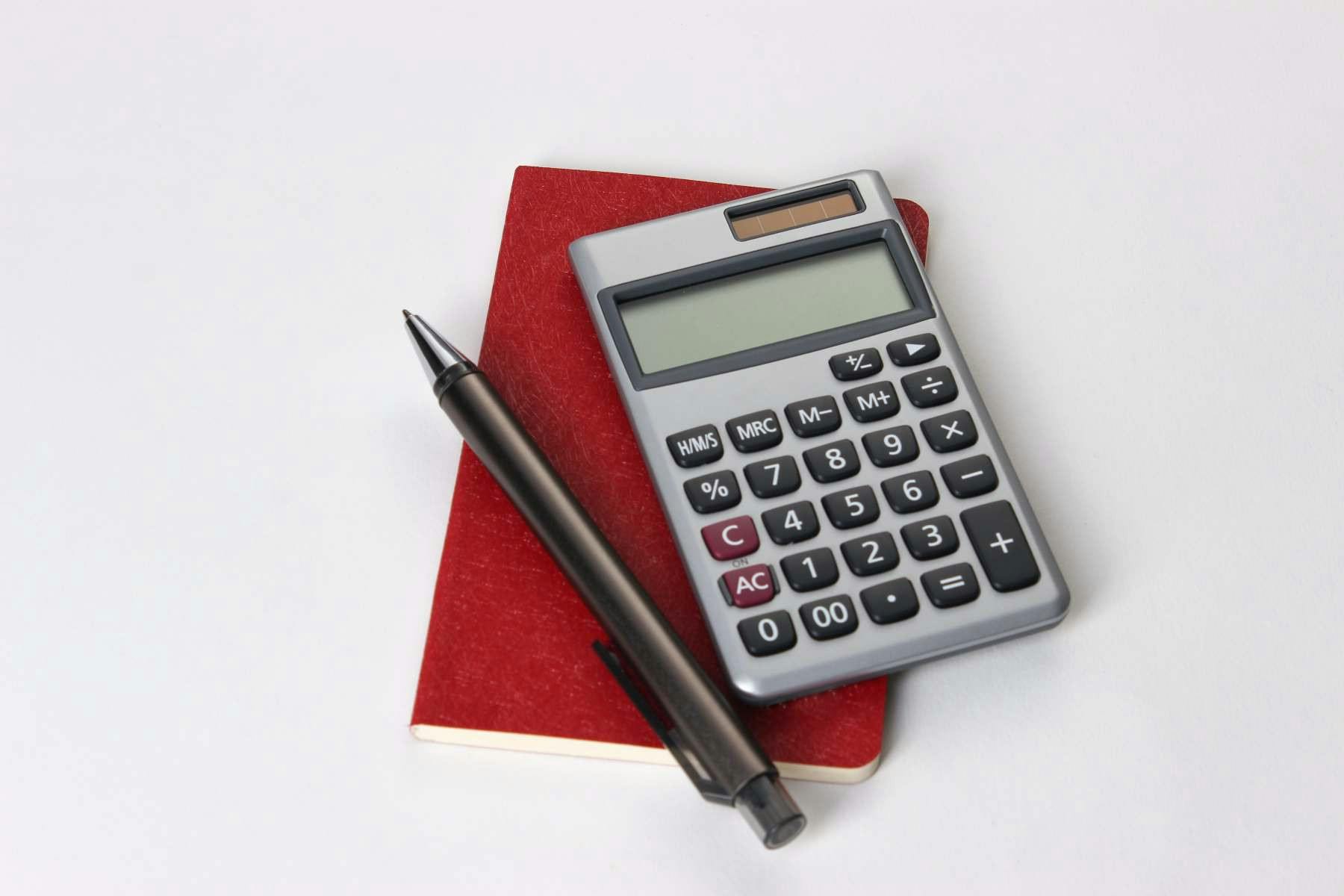 Imagem de uma calculadora, uma caneta preta e um caderno vermelho