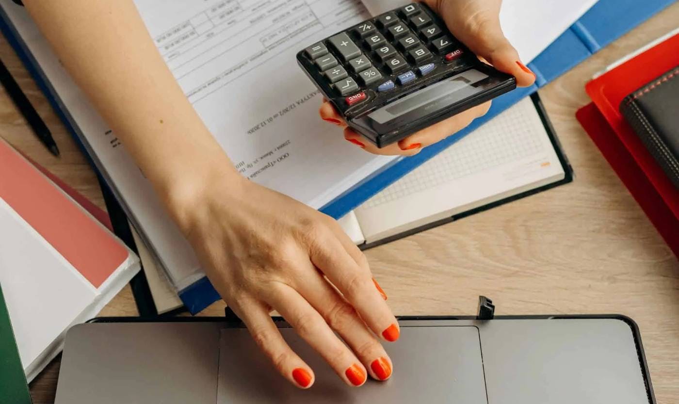 Imagem de uma mulher com esmalte vermelho. A foto está focada em suas duas mãos, uma delas segura uma calculadora e outra usa o mousepad de um notebook