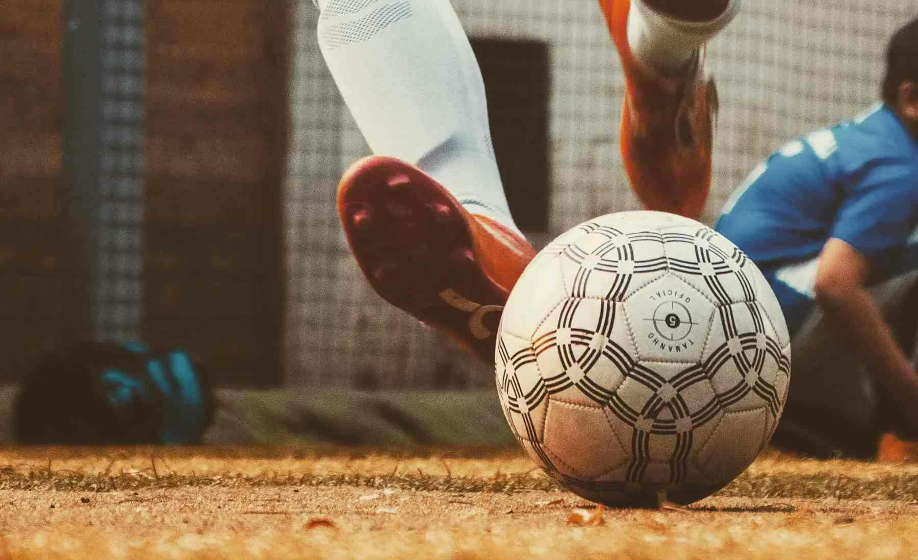 Imagem de uma bola de futebol em um campo, vemos também o pé de um atleta fazendo o chute. Ele usa chuteiras vermelhas
