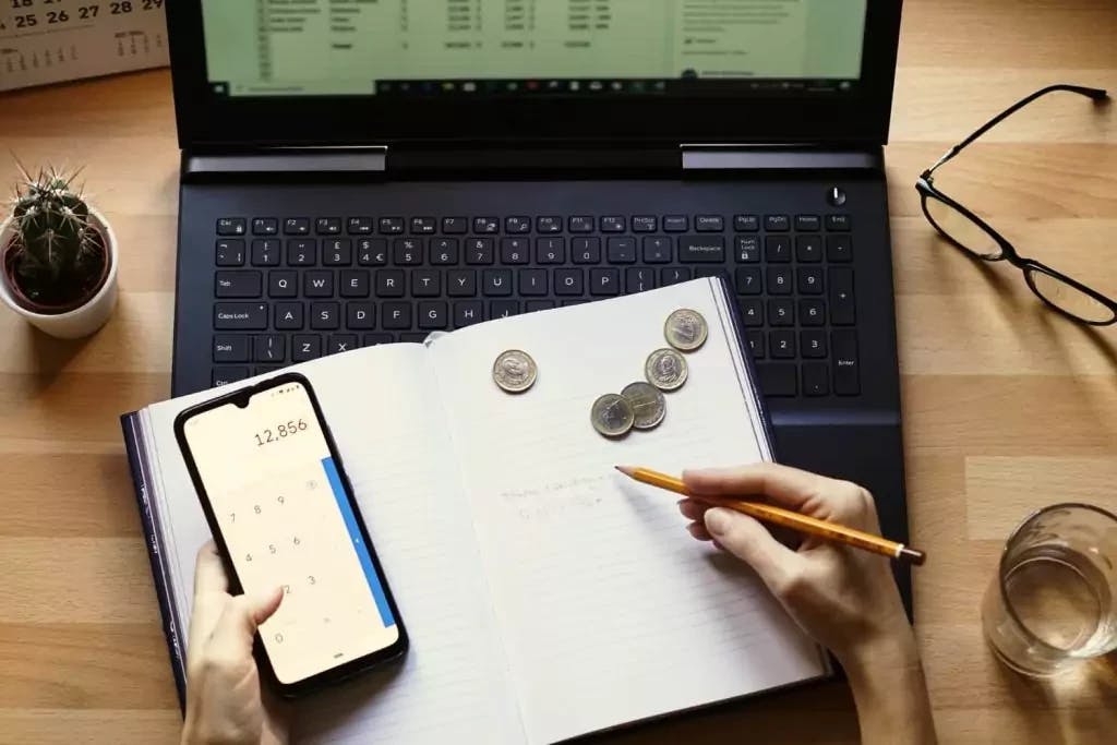 pessoa com papel e celular mostrando calculadora em cima de notebook