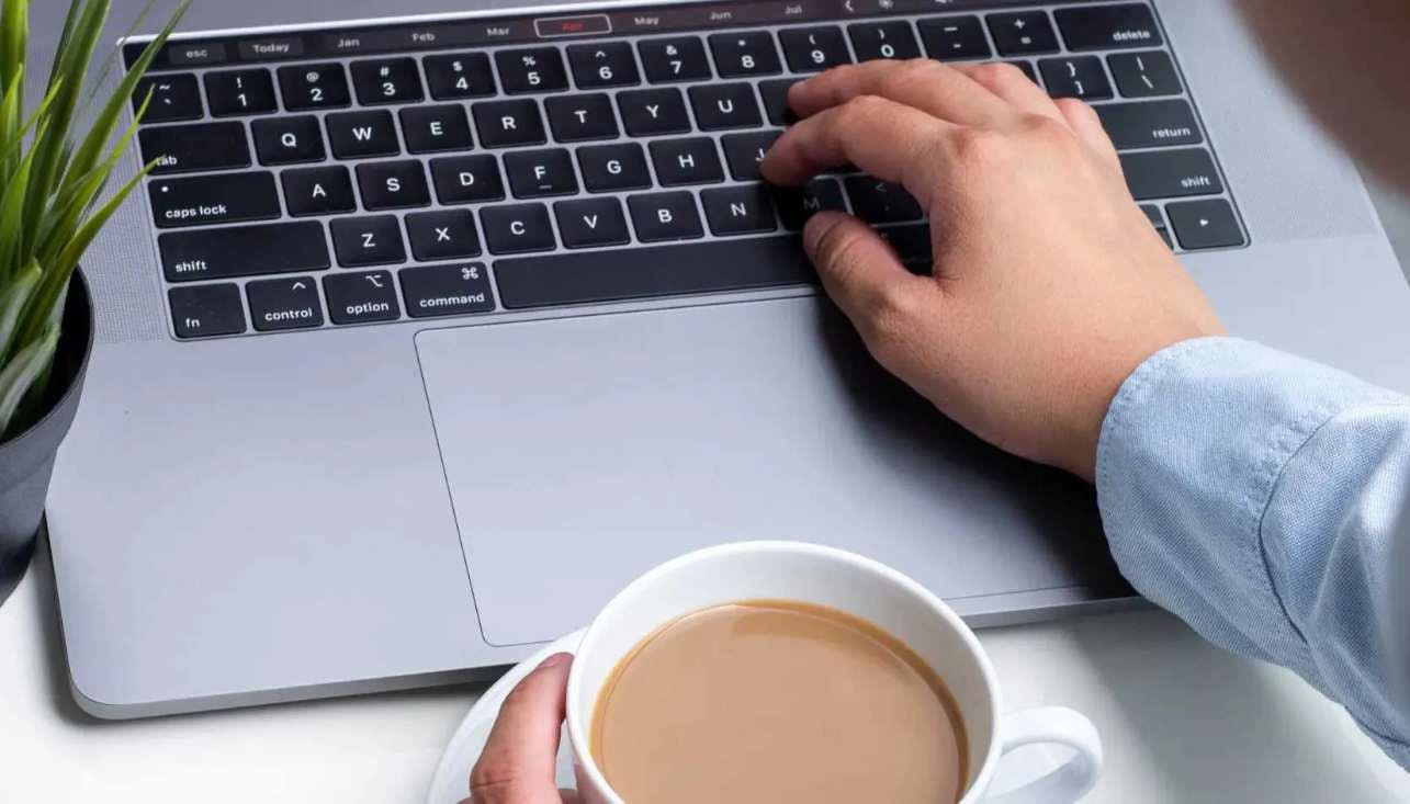 Imagem de um homem digitando no teclado de um notebook, com a outra mão ele segura uma xícara de café